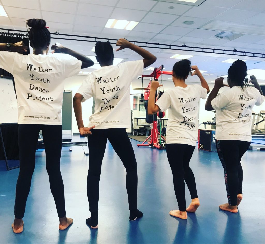 Four walker youth dance project participants. They are facing away from the camera, pointing at their shirts that read 'Walker Youth Dance Project' on the back.