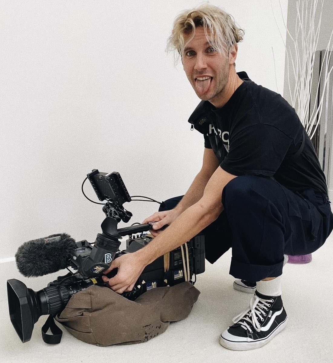 Jamie kneeling down with a camera in his hands while sticking his tongue out.