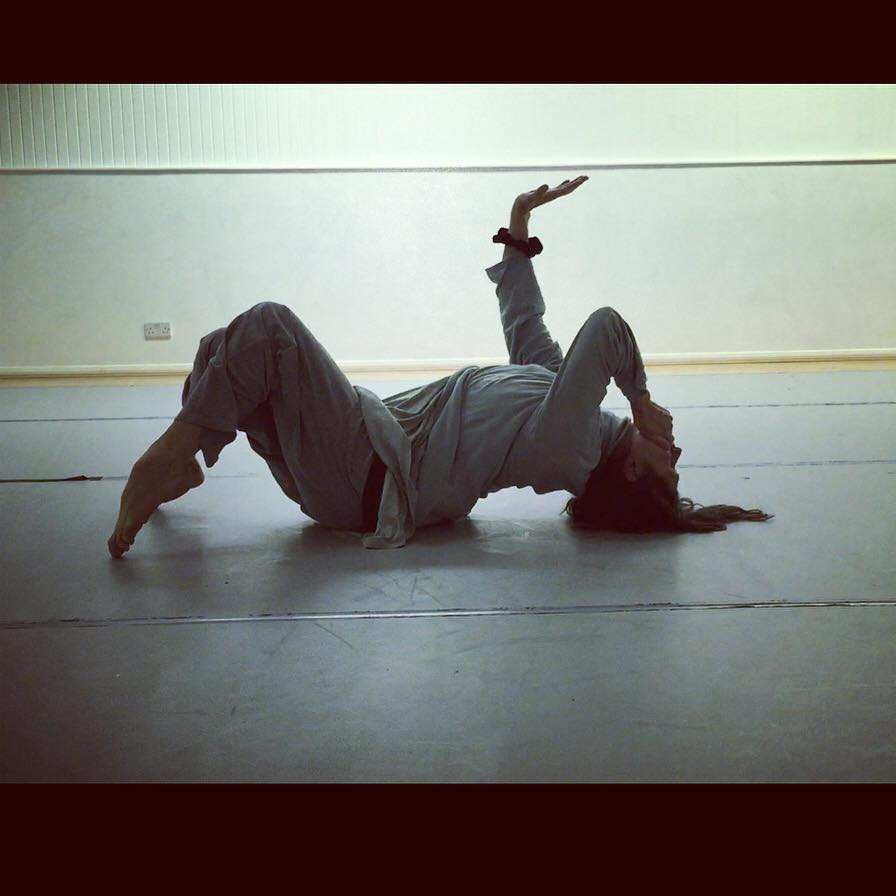 Caroline Reece dancing in rehearsal. She is lying on the floor on her back, with her back arched. One hand is over her mouth, with the other in the air, palm facing the sky. Her legs are bent and her toes are pointed, with only her toes on the floor.