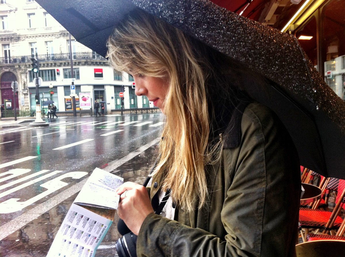 Pippa Howie facing left on a rainy street with an umbrella over her head. She is reading a flyer and wearing a green coat.