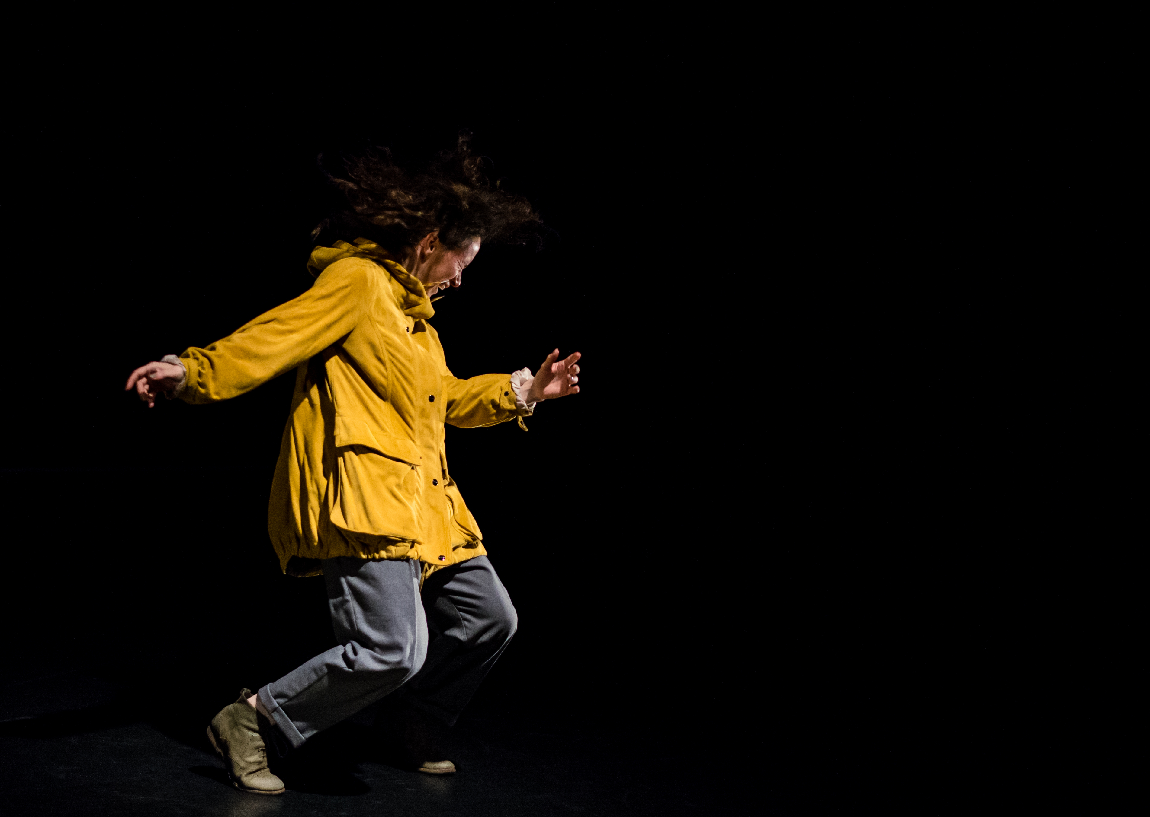 Kate dancing for The Sense of an Ending. Her legs are bent as she squats forward, her arms out. She is wearing a yellow coat, blue trousers and green shoes.