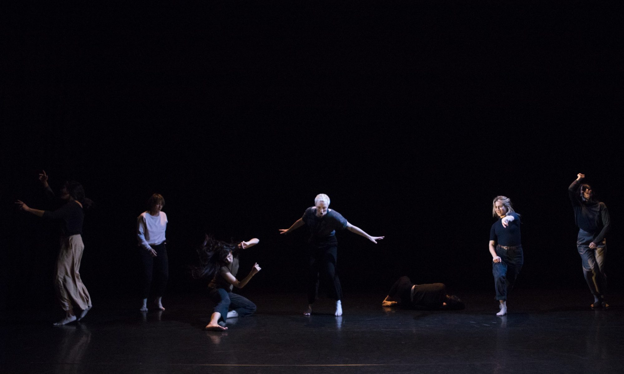 Seven dancers dancing in the dark. Left to right: A dancer facing stage left, turned away from the camera. They are mid jump and their arms are stretched out. A dancer standing facing towards the camera, stood slightly back on the stage. A dancer low on the floor, their left knee is on the floor and their right leg is stretched out towards the camera. Both their arms are bent and they are flicking their head towards the camera. A dancer stood in the centre of the stage, their arms are stretched out by their sides and they are looking at the floor. A dancer curled up on the stage floor, their back facing the camera and their head to the side. A dancer pointing towards the camera with their left hand, while stepping forwards. A dancer stood on the stage facing towards the camera, with their right arm above their head in a fist and the other by their legs.