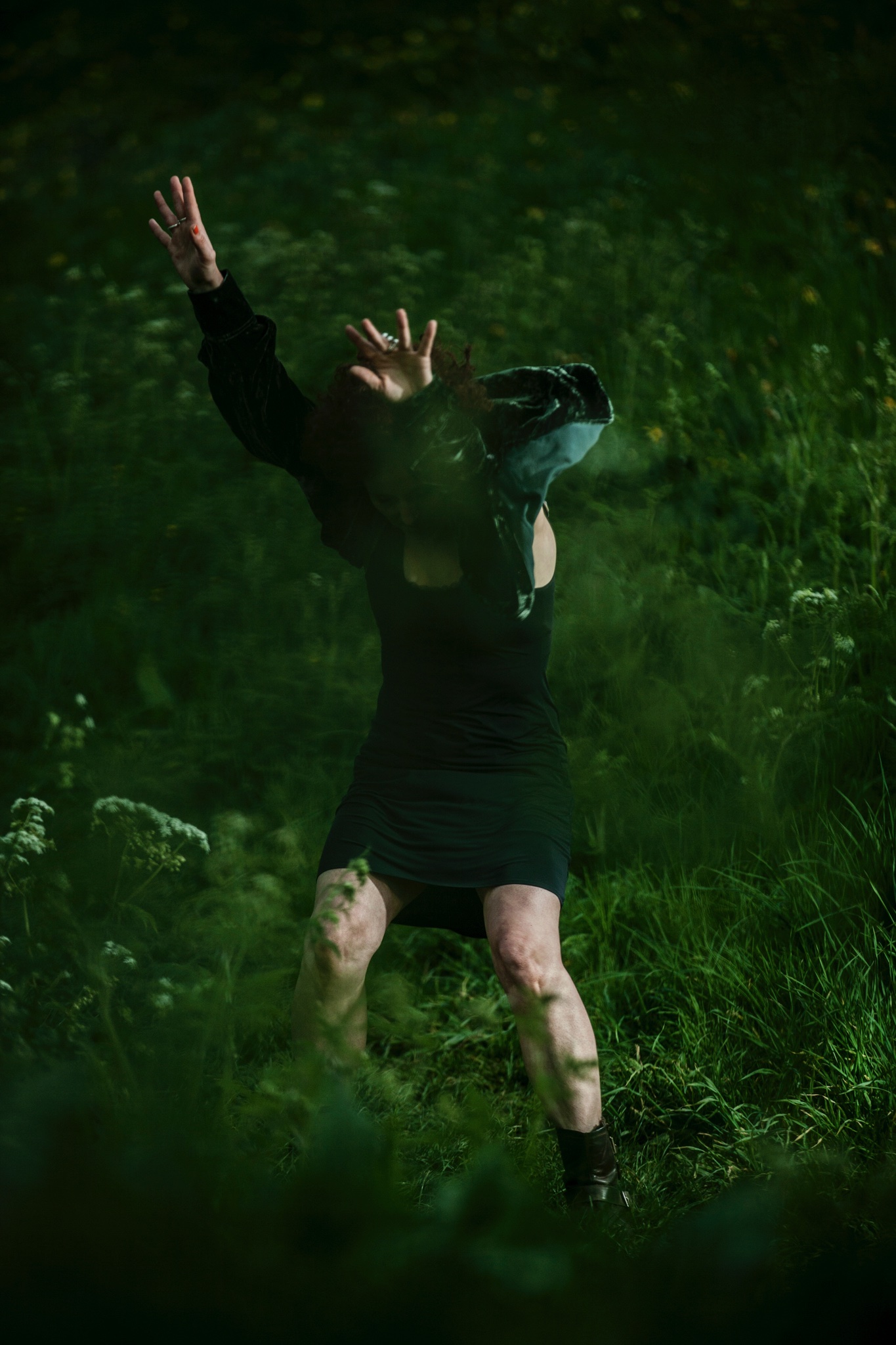 Keira Martin dancing in a completely green space. She is dancing in the grass, surrounded by bushes. Her legs are bent and her hands are above her head, hands outstretched, with a shawl she is wearing covering her face.