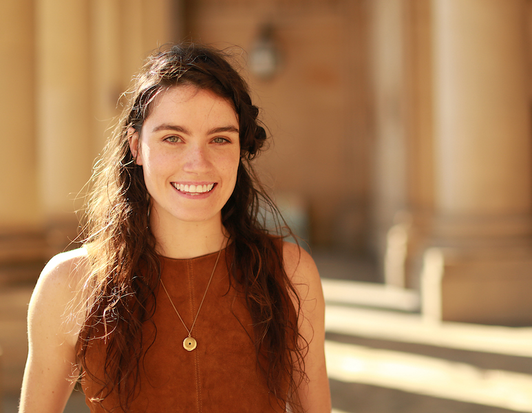 Headshot of Sophie Hutchinson stood outside. She is smiling wide and facing the camera, wearing a brown sleeveless dress and long necklace.