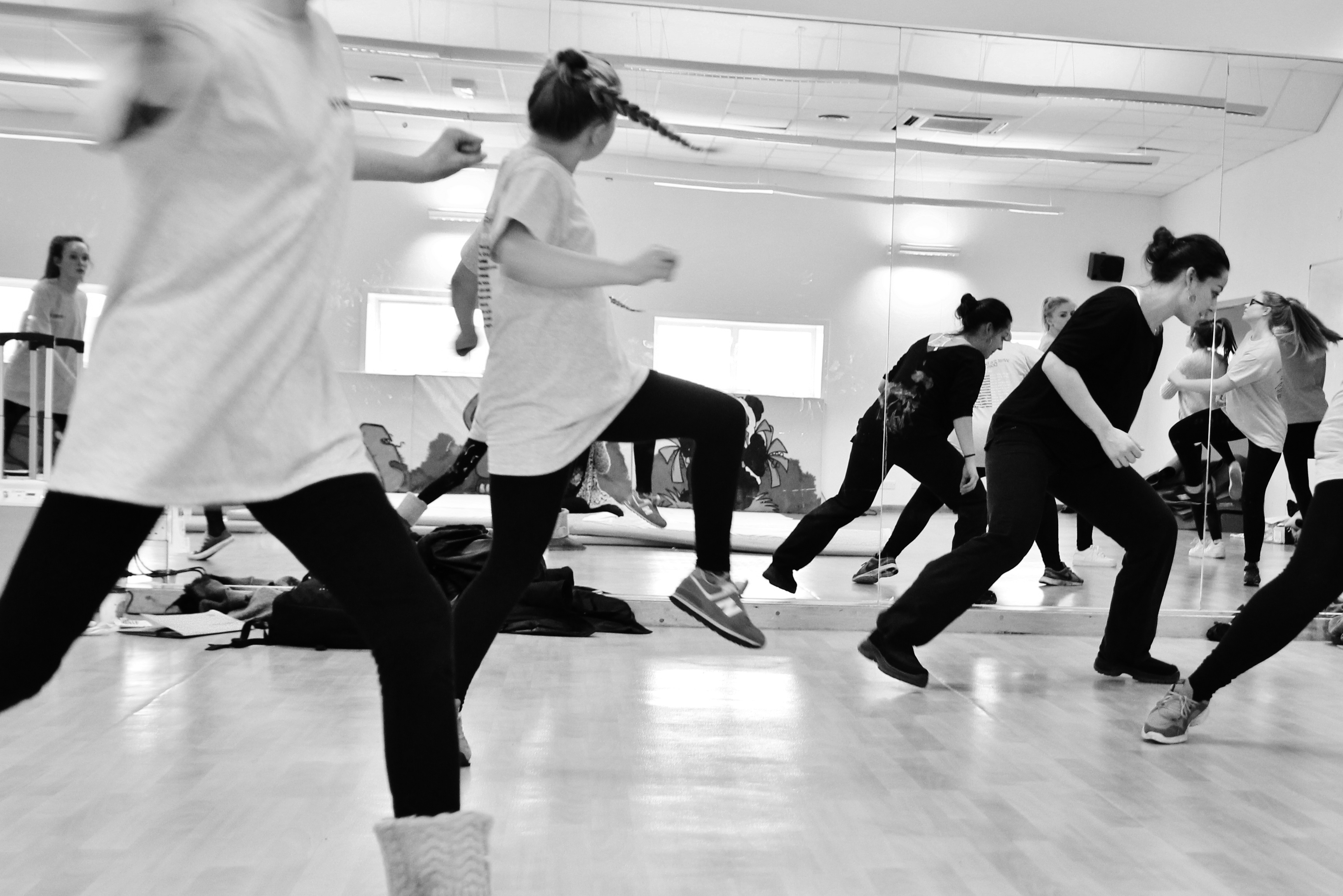 Nadia Iftkhar running a youth dance class. In the foreground, there are two dancers jumping, with their legs bent, spinning. Nadia is in the background, lunging forward. They are dancing in front of a studio mirror, in which you can see more dancers rehearsing.