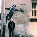 Megan Brown outside, stood in front of streetart by artist Kuba, depicting Gene Kelly dancing in Singing in the Rain. She is smiling and tucking her hands into her pockets.