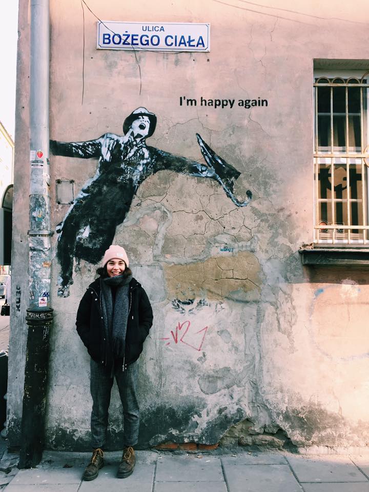 Megan Brown outside, stood in front of streetart by artist Kuba, depicting Gene Kelly dancing in Singing in the Rain. She is smiling and tucking her hands into her pockets.