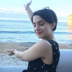 Elif Emma True on the beach, smiling with her eyes closed. Her right arm is stretched straight up in the air and her left arm stretched out in front of her body, as if presenting the sea.