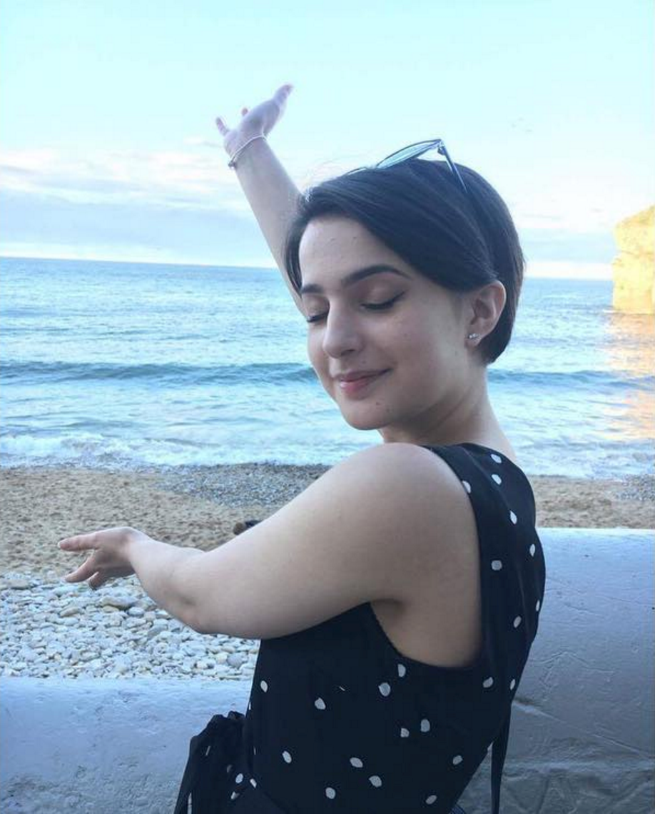 Elif Emma True on the beach, smiling with her eyes closed. Her right arm is stretched straight up in the air and her left arm stretched out in front of her body, as if presenting the sea.