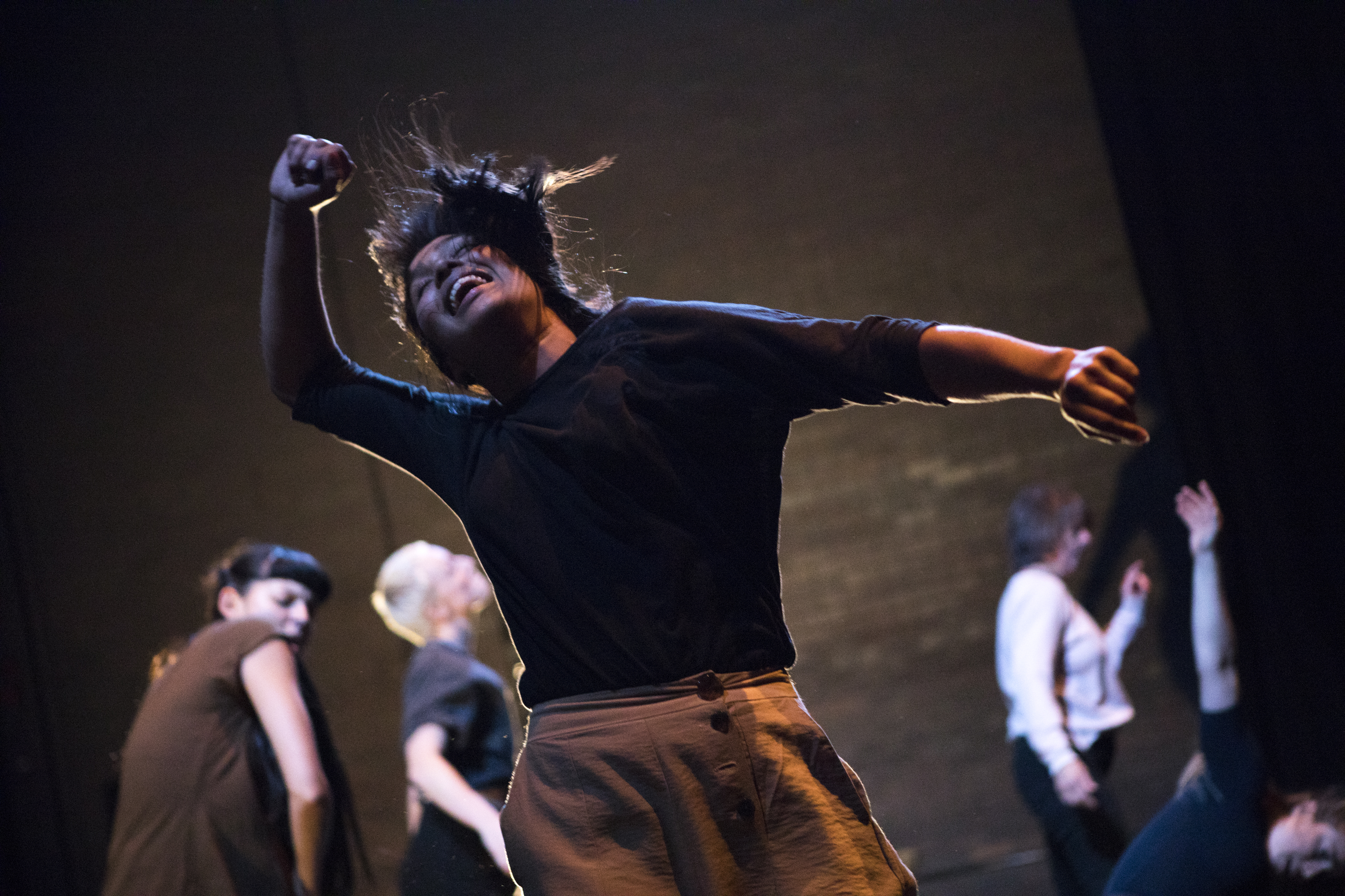 Performers dancing for Crackle Dust. In the centre of the image, a dancer is mid movement, hair in the air, with her arms out to the side. There are four dancers in the background, all mid movement, all facing stage right. The dancer on the right is dipping low, their their arm straight up in the air.