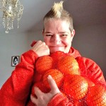 Al Orange smiling at the camera. She has a mohawk, and is holding a big bag of oranges up to her chest. She is in a room with a chandelier and a framed piece of artwork behind her.