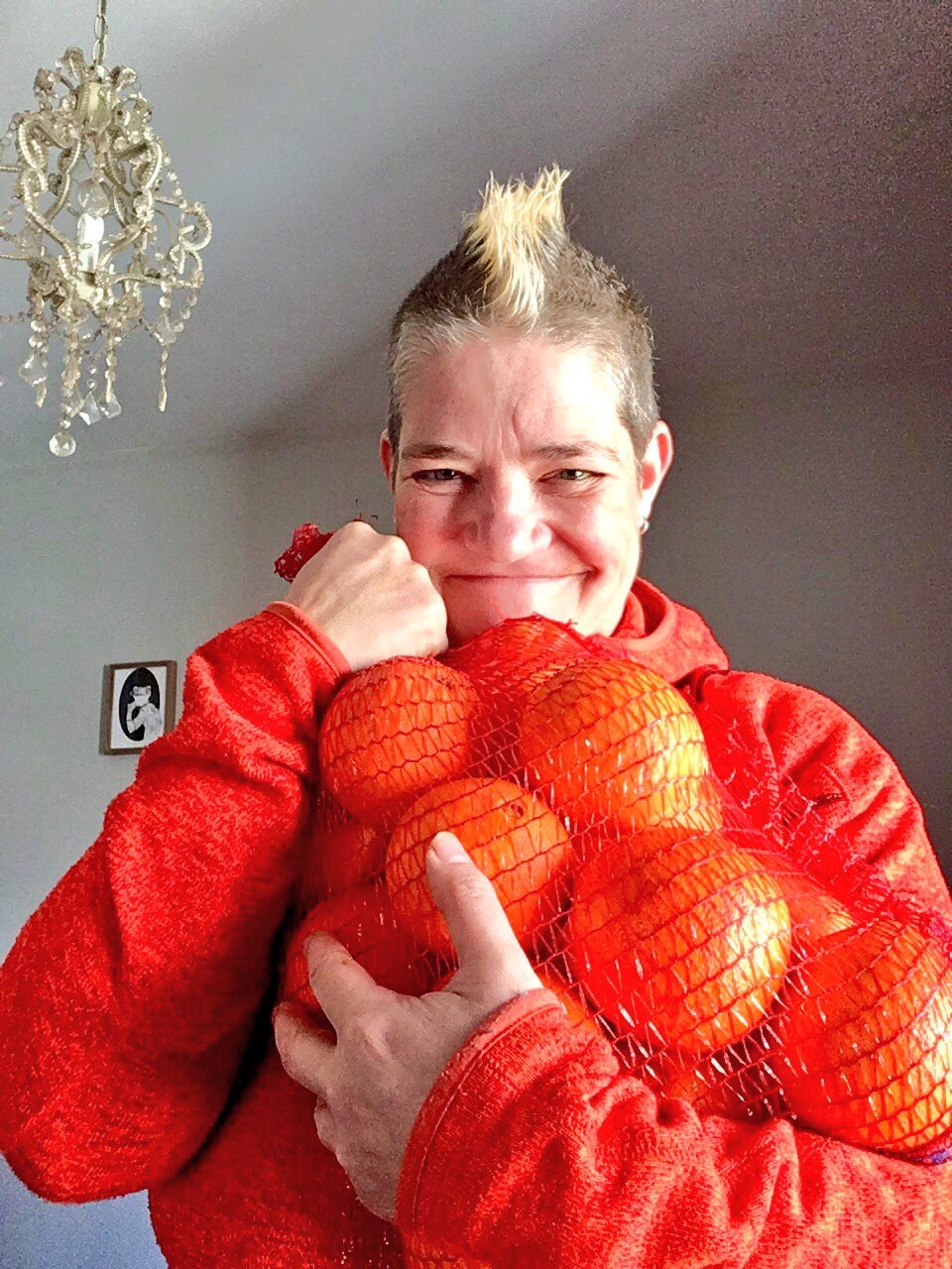 Al Orange smiling at the camera. She has a mohawk, and is holding a big bag of oranges up to her chest. She is in a room with a chandelier and a framed piece of artwork behind her.