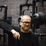 Nicolai Hart-Handsen leaning on a stair railing. Behind him is theatre technical equipment. He is wearing big, thick rimmed glasses and has a short beard.