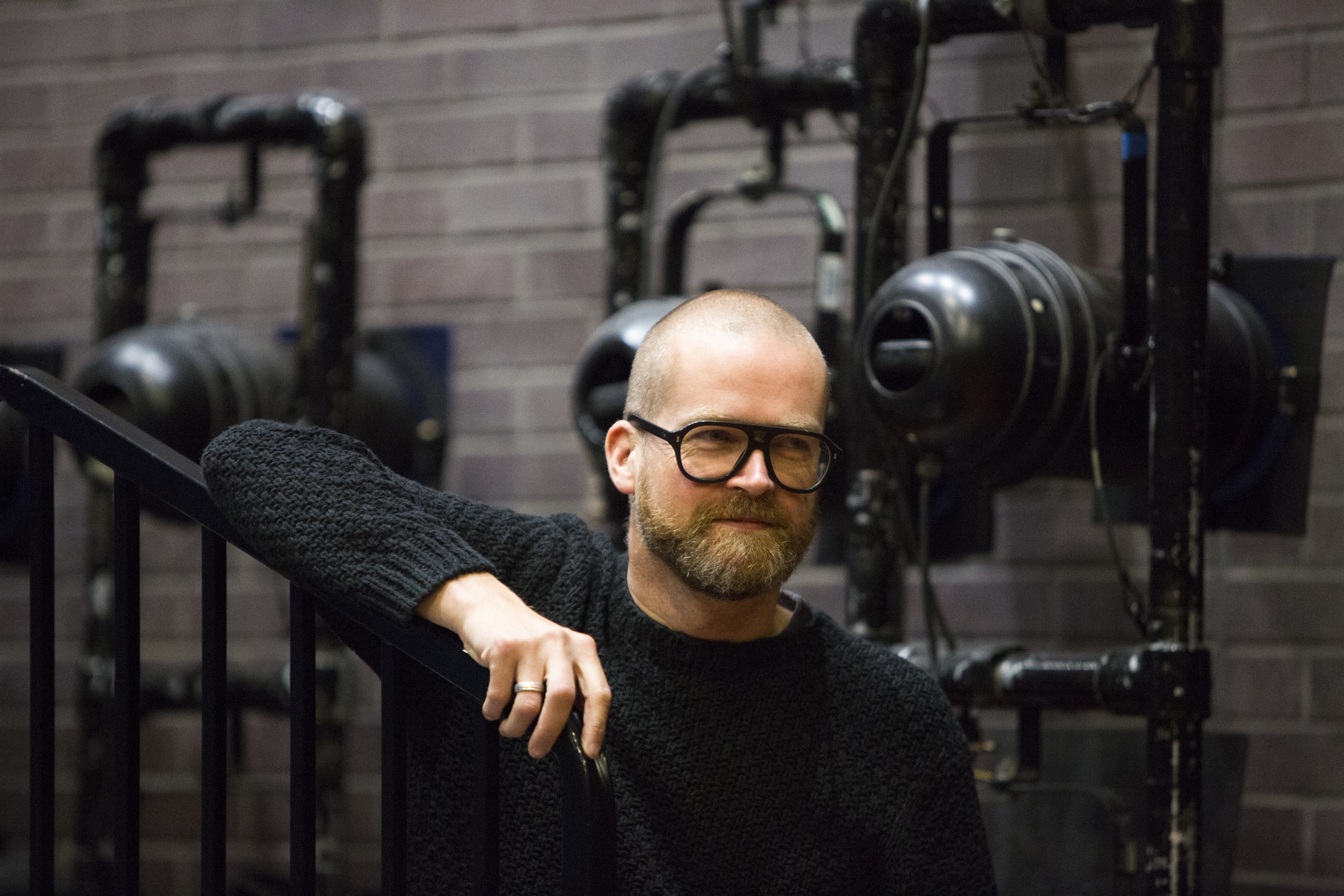 Nicolai Hart-Handsen leaning on a stair railing. Behind him is theatre technical equipment. He is wearing big, thick rimmed glasses and has a short beard.