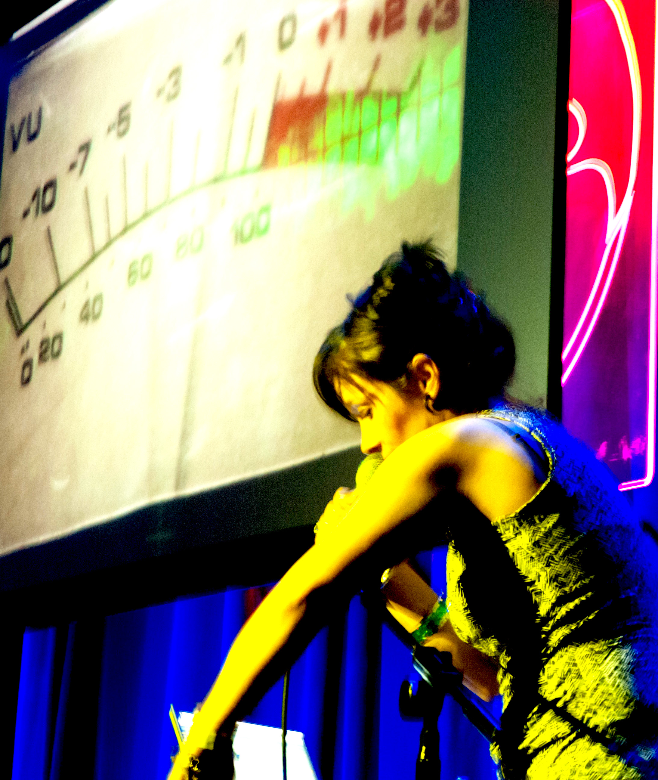 Caro C performing on stage. She is singing into a microphone she’s holding in her right hand. She is lit with a yellow light and is stood in front of a screen with a metre projected onto it, a neon light and a stage curtain.