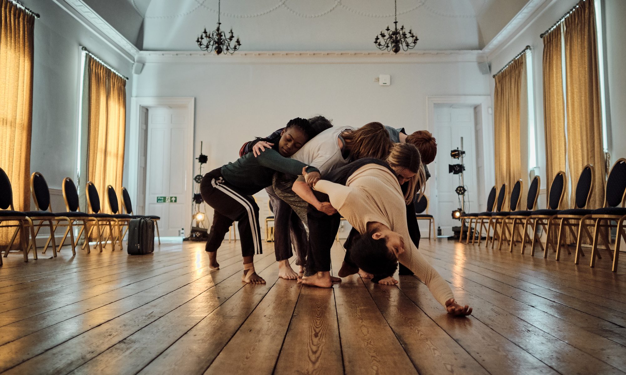 Six dancers rehearsing for HELD 2021 in a large room with wooden floors and chandeliers. One dancer is falling back with their right hand resting on the floor as the other five dancers support them by holding them, stopping them from falling, as they rest their heads on the dancer and eachtother.