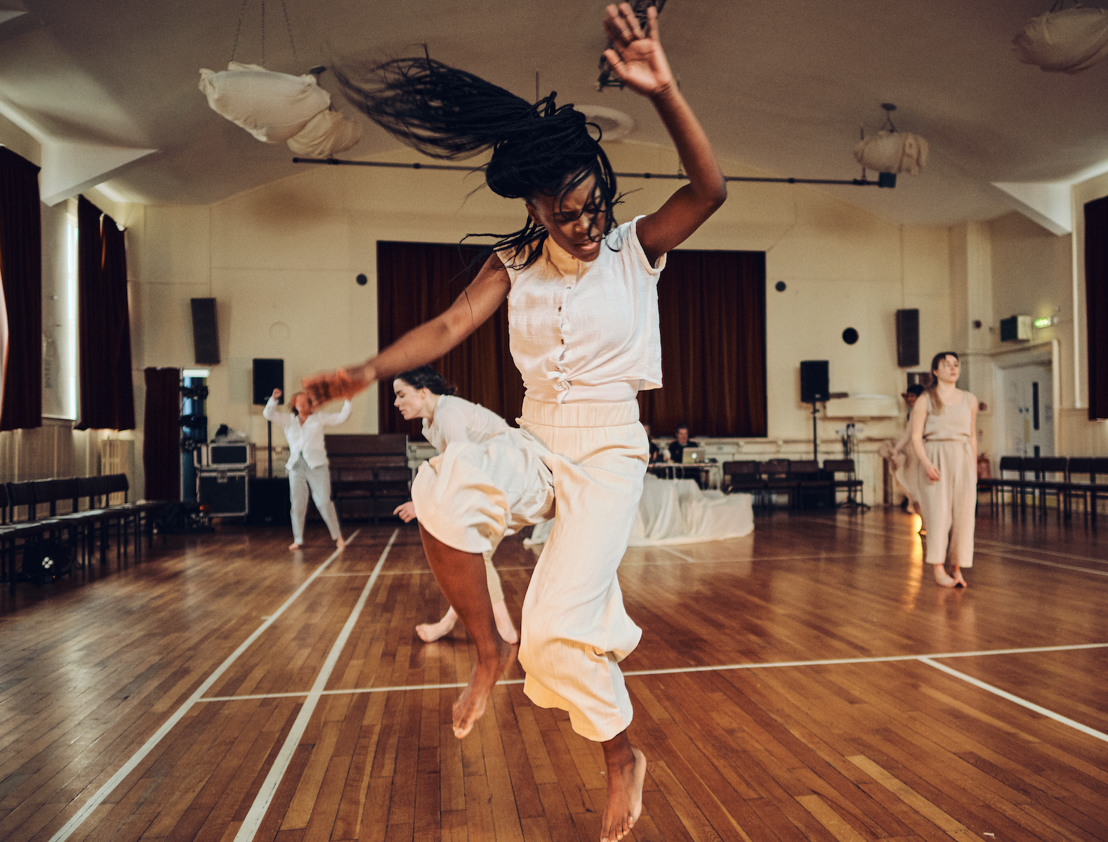 Benny Valentina jumping during a rehearsal. Her hair is flying in the air, with her right arm up towards the sky, the other reaching out to her side. Behind her are three other dancers rehearsing.