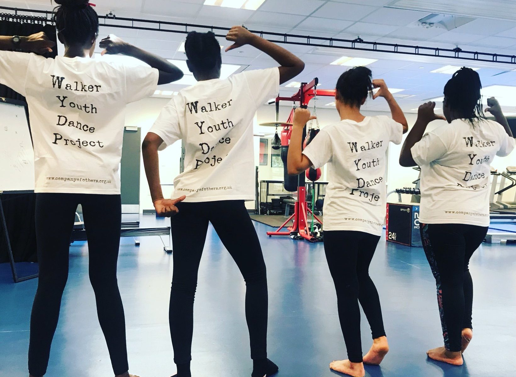Four walker youth dance project participants. They are facing away from the camera, pointing at their shirts that read 'Walker Youth Dance Project' on the back.