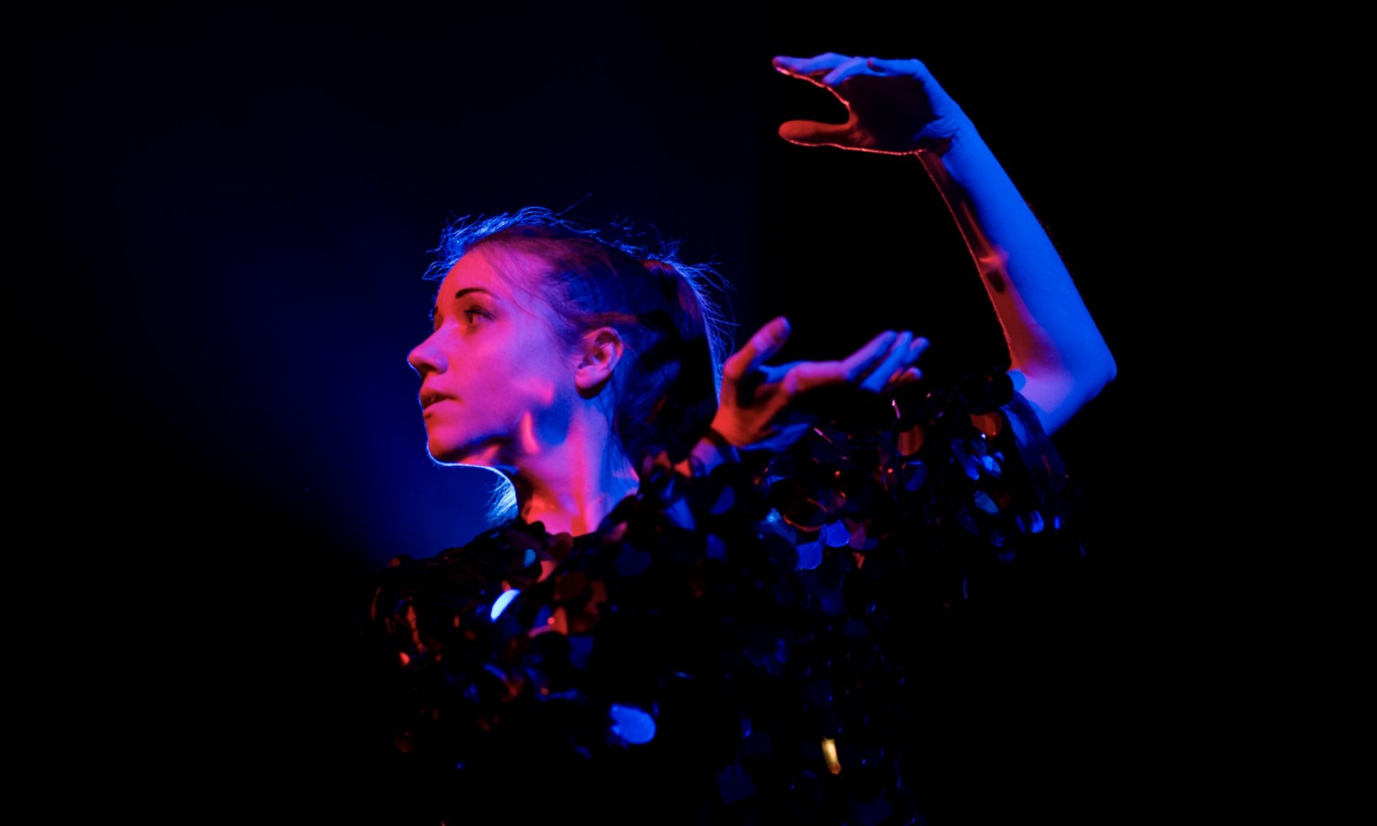 Jem Clancy dancing on stage. Her arm is up high behind her. She is lit by purple, pink and blue lighting.