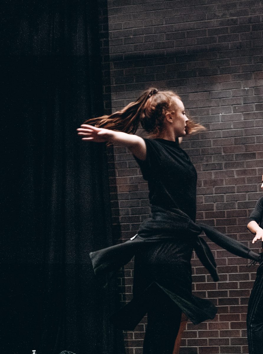 Cora-Jay Williams dancing in rehearsal. She is spinning, with her arms stretched out wide. Her hair and the hoody she has tied around her waist are in the air from the momentum.