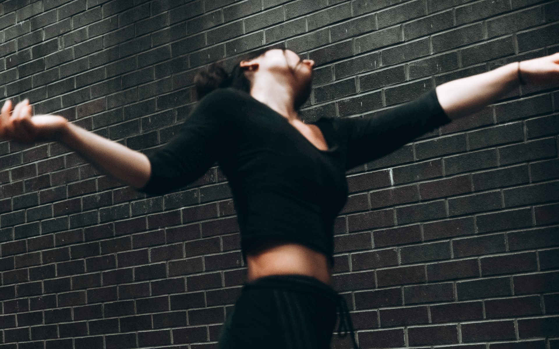 Lila from a low angle, dancing in front of a dark brick wall. Her arms are stretched out to her side, her head turned away from the camera.