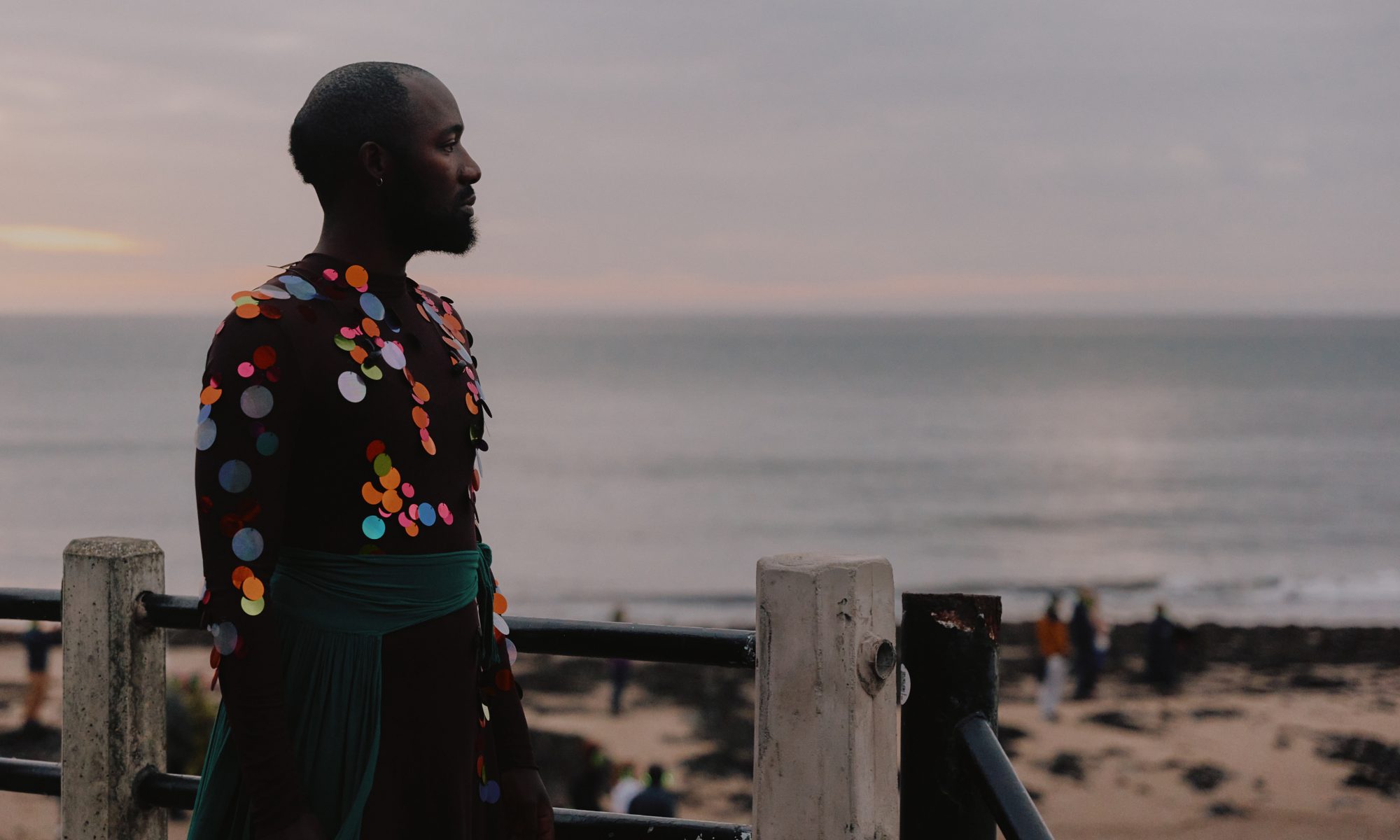 Patrick Ziza dressed in a mesh and sequin outfit standing by a railing looking at the ocean.