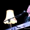 Beccy Owen performing on stage. She is playing a keyboard, singing into a microphone. Behind her is a lamp that is turned on.