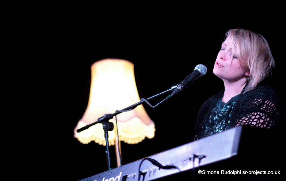 Beccy Owen performing on stage. She is playing a keyboard, singing into a microphone. Behind her is a lamp that is turned on.