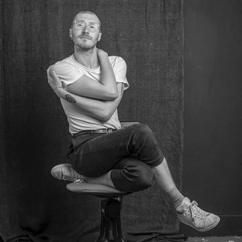 Black and white portrait of Phillip Brown sat on a stool. One leg is bent underneath him with the other crossed on top of it. He's holding his body with his arms.