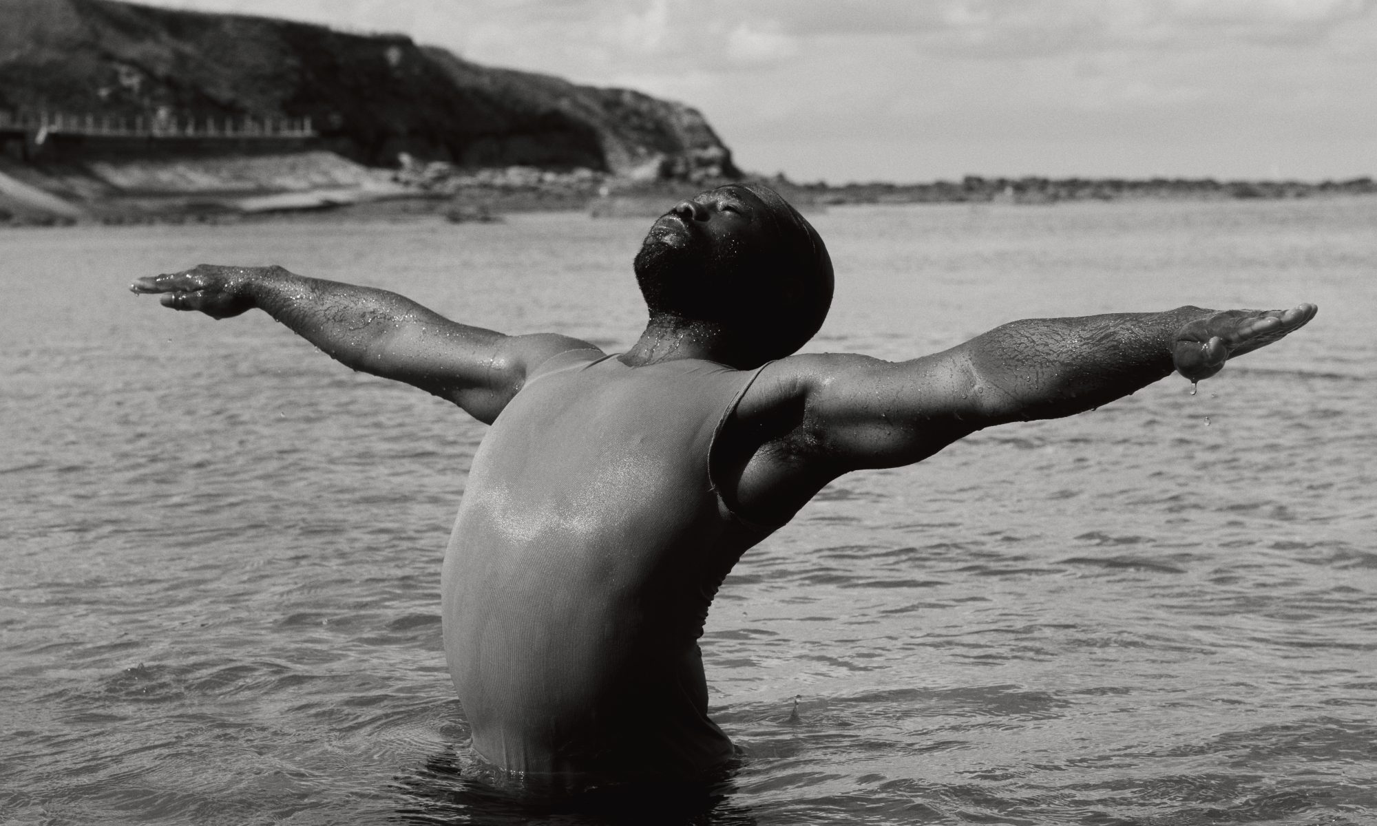 Jeremiah standing waist deep in the ocean, leaning backwards, arms spread out on each side while facing the sky with his eyes closed.