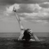 Amy Becke in the ocean looking to the sky with her left arm raised in the air and right hand placed on her head.