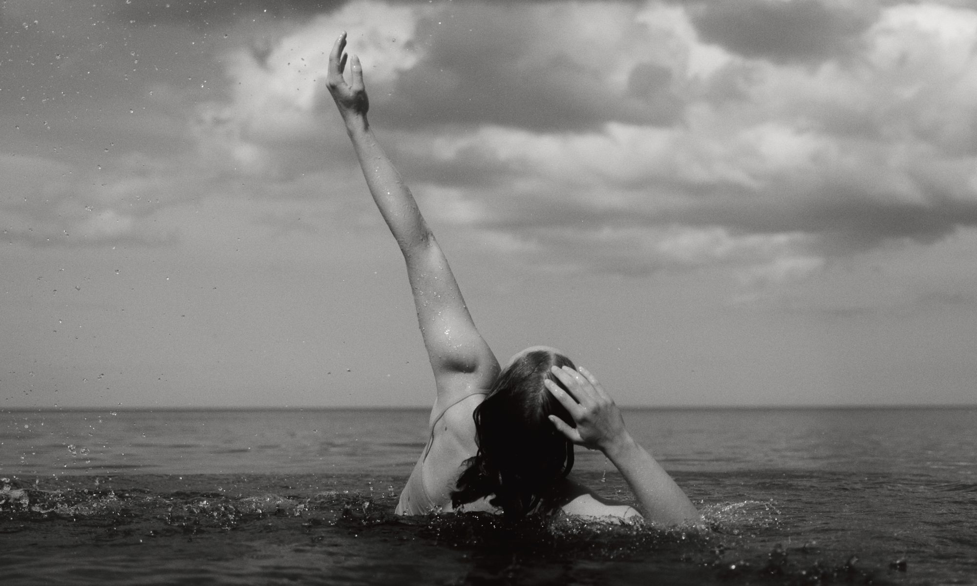 Amy Becke in the ocean looking to the sky with her left arm raised in the air and right hand placed on her head.