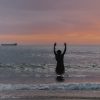 Amari standing knee deep in the ocean with his hands raised facing the sunset.