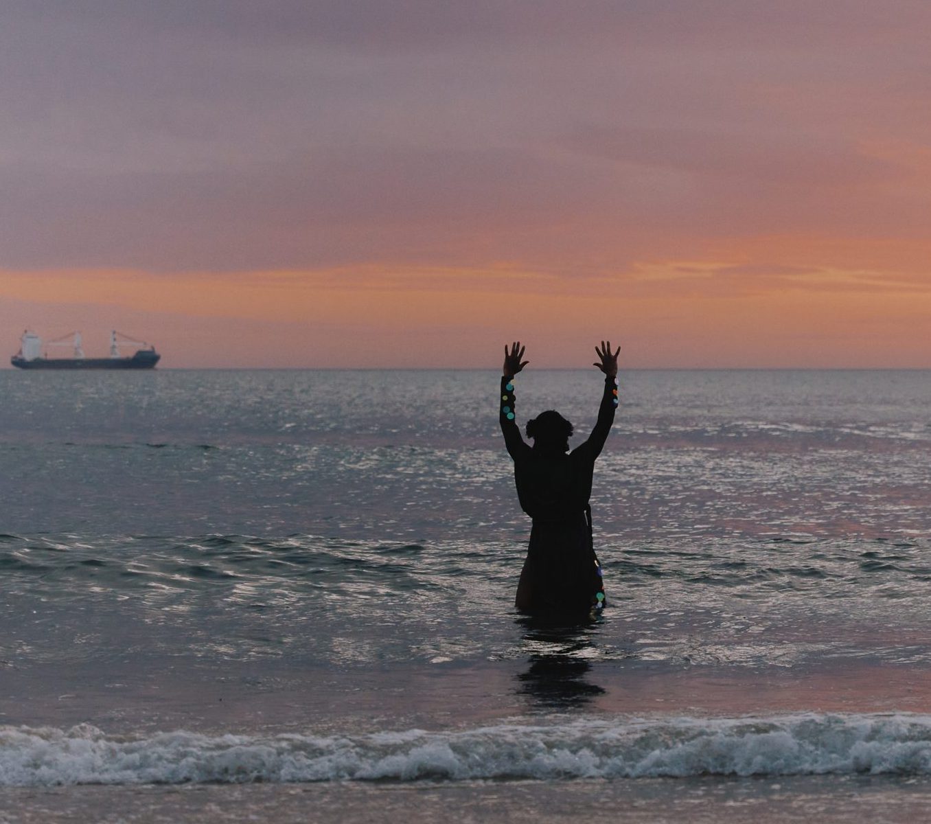 Amari standing knee deep in the ocean with his hands raised facing the sunset.