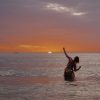 Michaela Wate standing in the ocean at sunset with her left arm raised towards the sky and right arm bent on her side.
