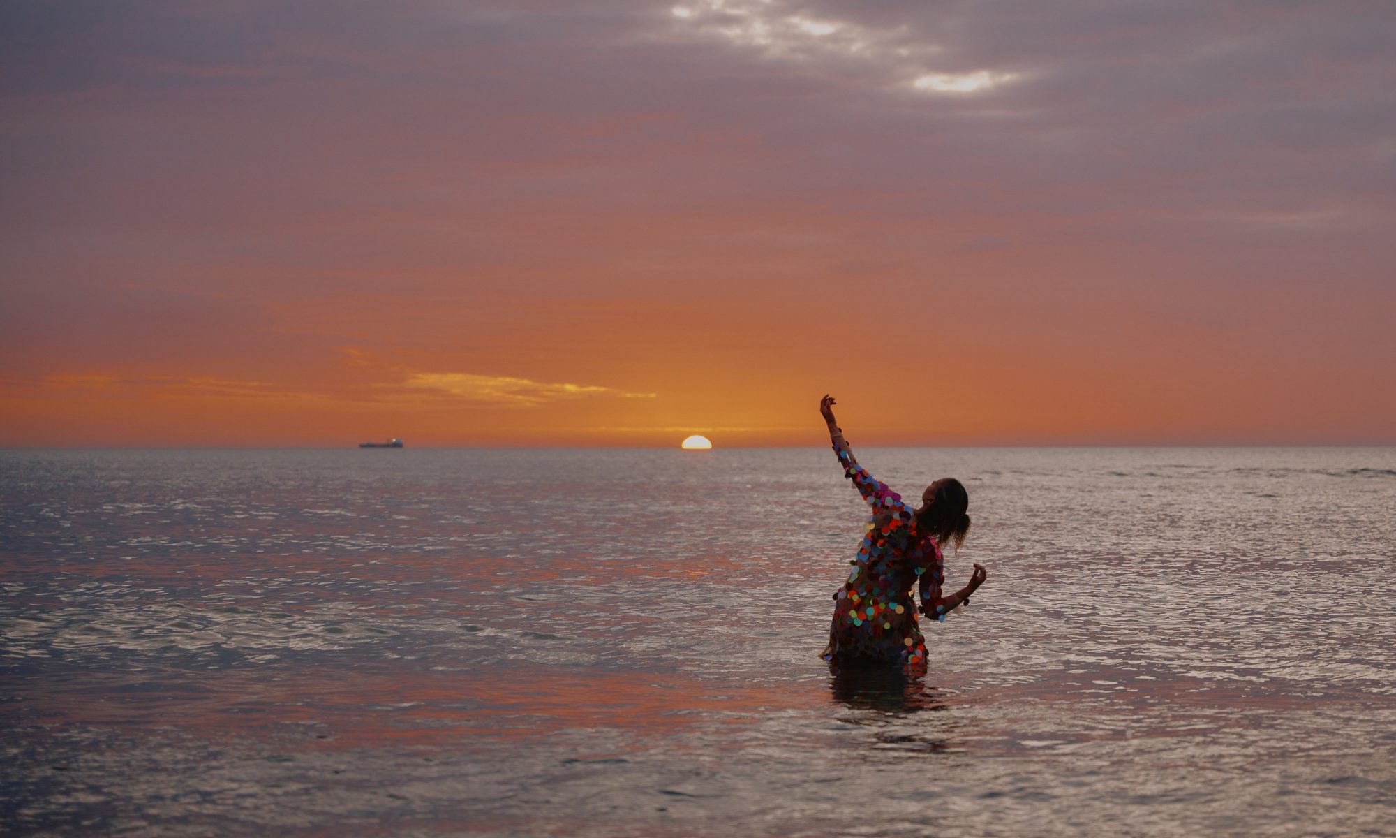 Michaela Wate standing in the ocean at sunset with her left arm raised towards the sky and right arm bent on her side.