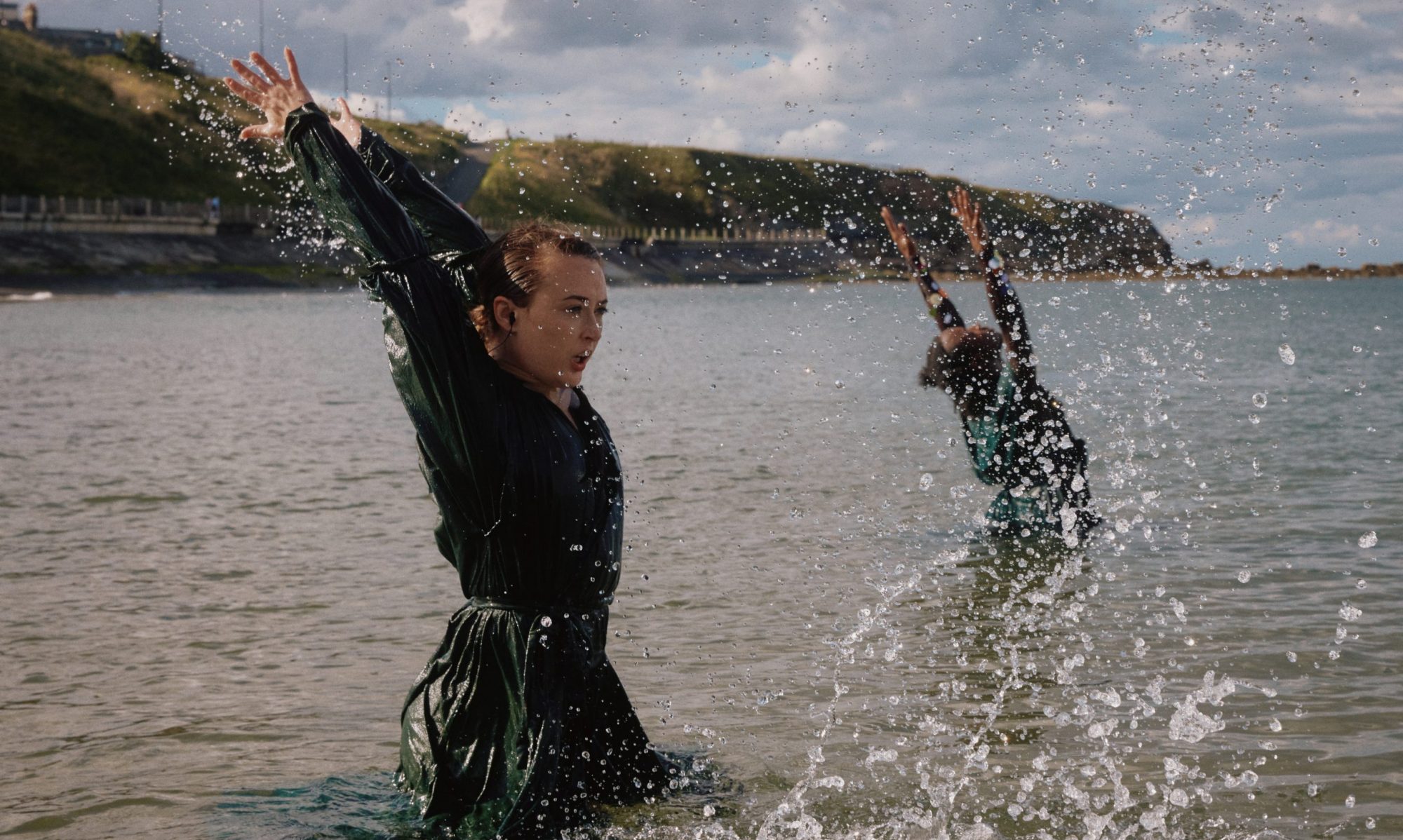 Two dancers standing waist deep in the ocean. Their arms are raised and backs arched as water splashes around them.