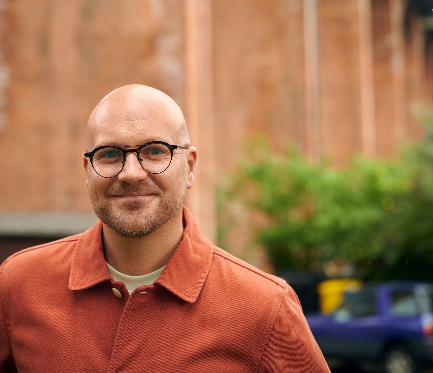 A person wearing glasses and an orange jacket smiles warmly in front of an old brick viaduct. The scene conveys a relaxed, urban vibe.