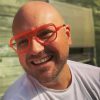 A portrait of Gareth smiling. He has on a whit shirt and a pair of glasses with red frames.