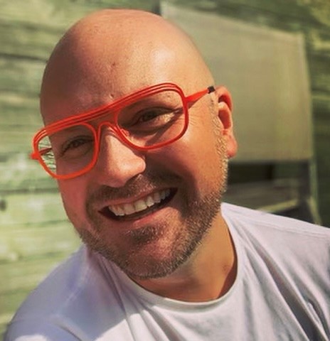 A portrait of Gareth smiling. He has on a whit shirt and a pair of glasses with red frames.