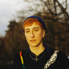 Em pictured outdoors against some trees. They have short orange hair, and wear a beanie and jumper.