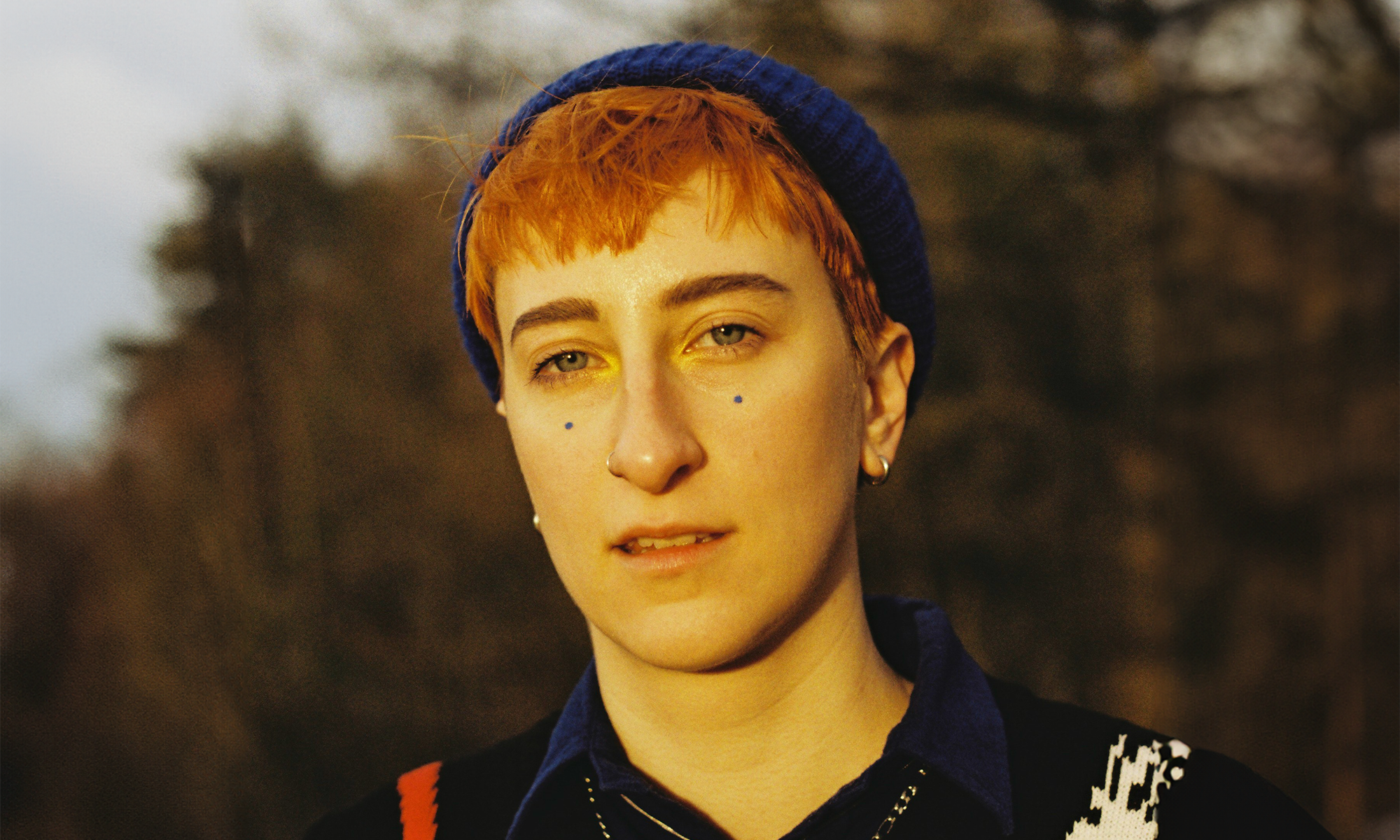 Em pictured outdoors against some trees. They have short orange hair, and wear a beanie and jumper.
