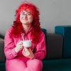 Person with curly red hair in a pink outfit sits on a bench, smiling warmly while holding a white coffee cup. The mood is cheerful and relaxed.