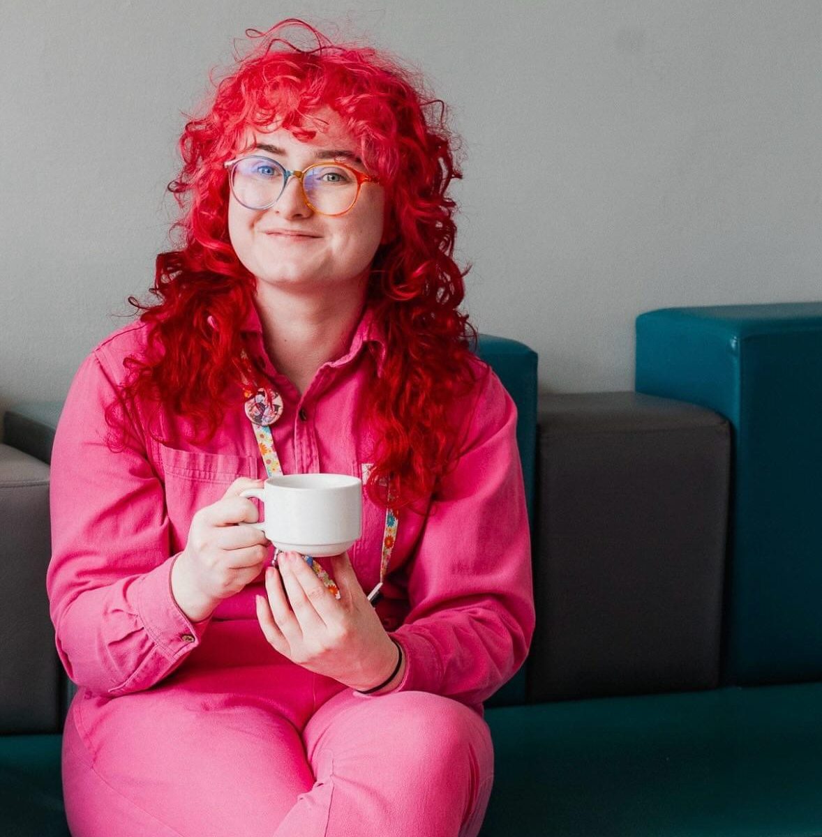 Person with curly red hair in a pink outfit sits on a bench, smiling warmly while holding a white coffee cup. The mood is cheerful and relaxed.